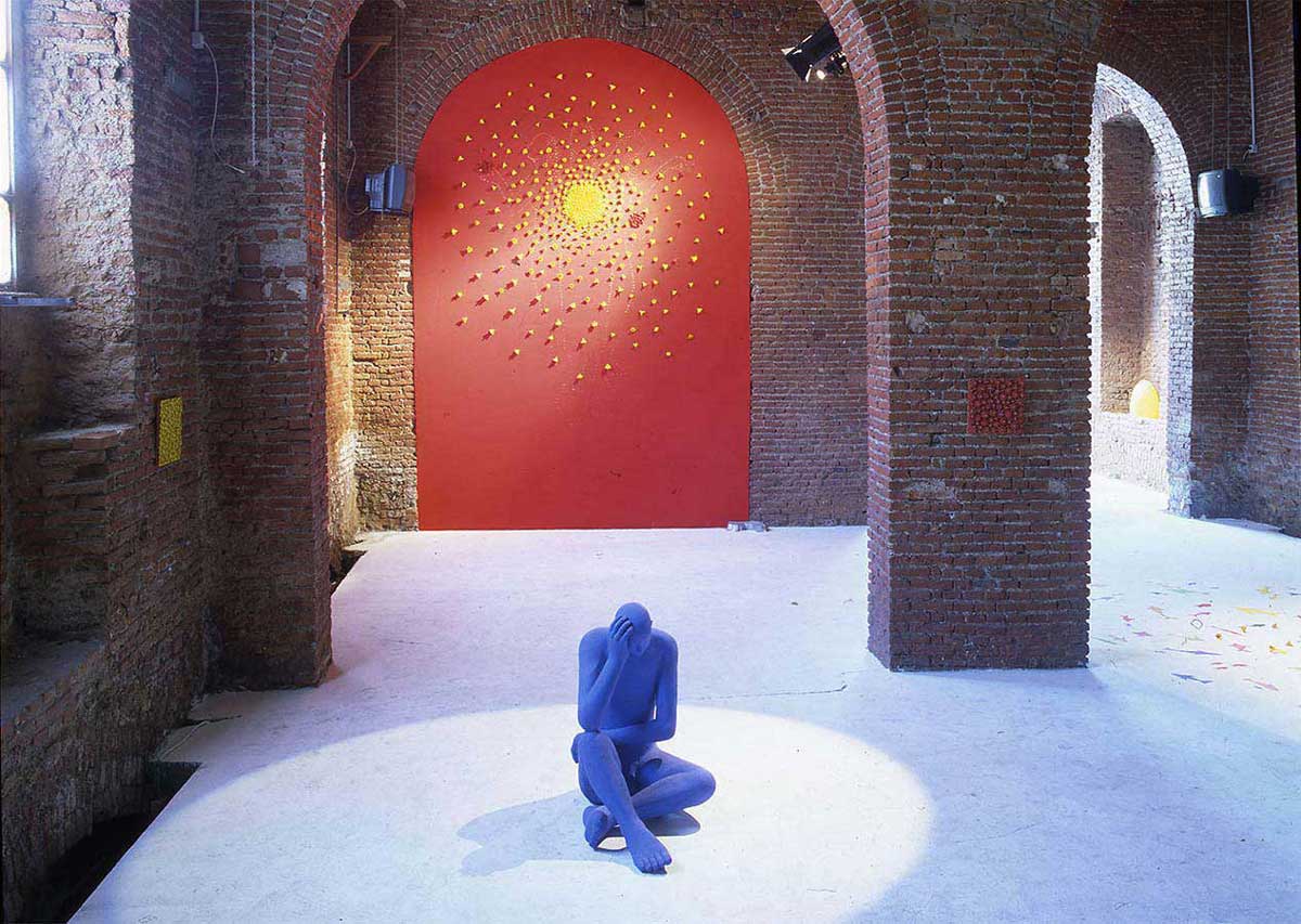 The scene of the Kazumasa Mizokami exhibition held at the EOS Factory in Milan in 1998. A blue seated statue in the foreground and a yellow flower on the large red wall at the back.  photo by studio Acqua.