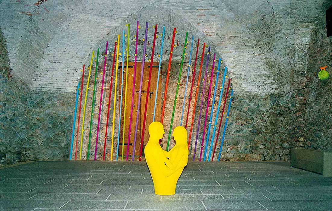 2004 A  exhibition of Kazumasa Mizokami, in the church of San Agostino in Pietrasanta. Using the curve of the building on the back, the colored bars are aligned to make it look like a rainbow. There is a sculpture in front of which yellow men and women embrace