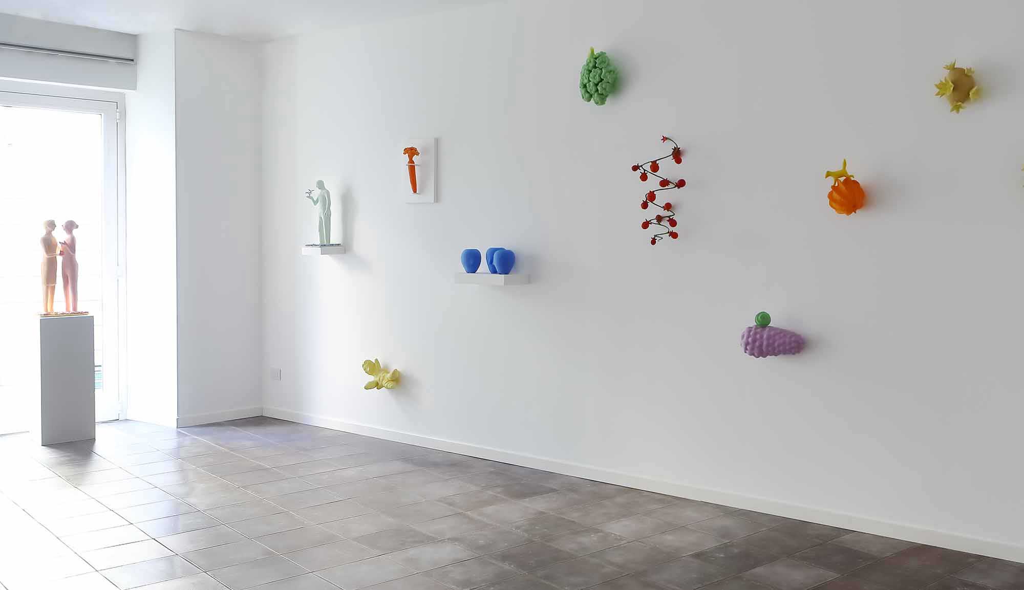 Exhibitions of gallery melisi 2015 A large white wall features 9 small multicolored vegetable sculptures by Kazumasa Mizokami. In the far left corner there is a pink statue of a little friend.