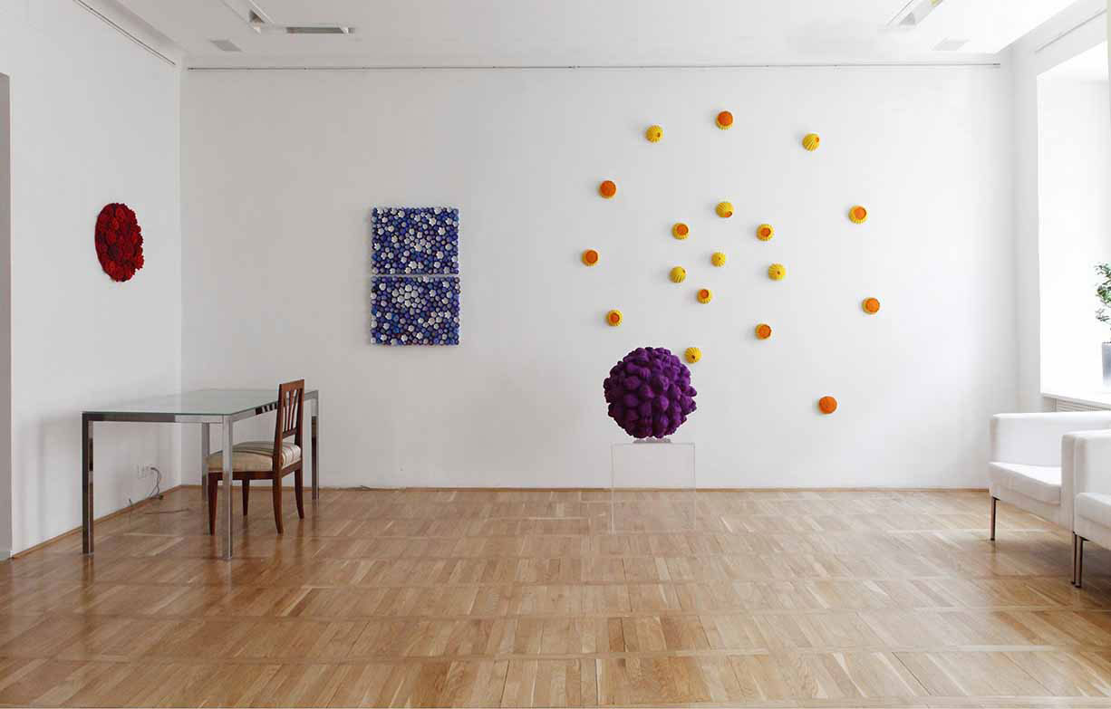Shot of exhibitions-gallery-visconti 2011 On the white wall of the exhibition hall, there are 18 small hemispherical sunflower-shaped objects painted in yellow and orange scattered in the shape of a spiral. On the wall, a rectangular white blue work. A purple surface is worked in front of them. a purple sphere sculpture by Kazumasa Mizokami