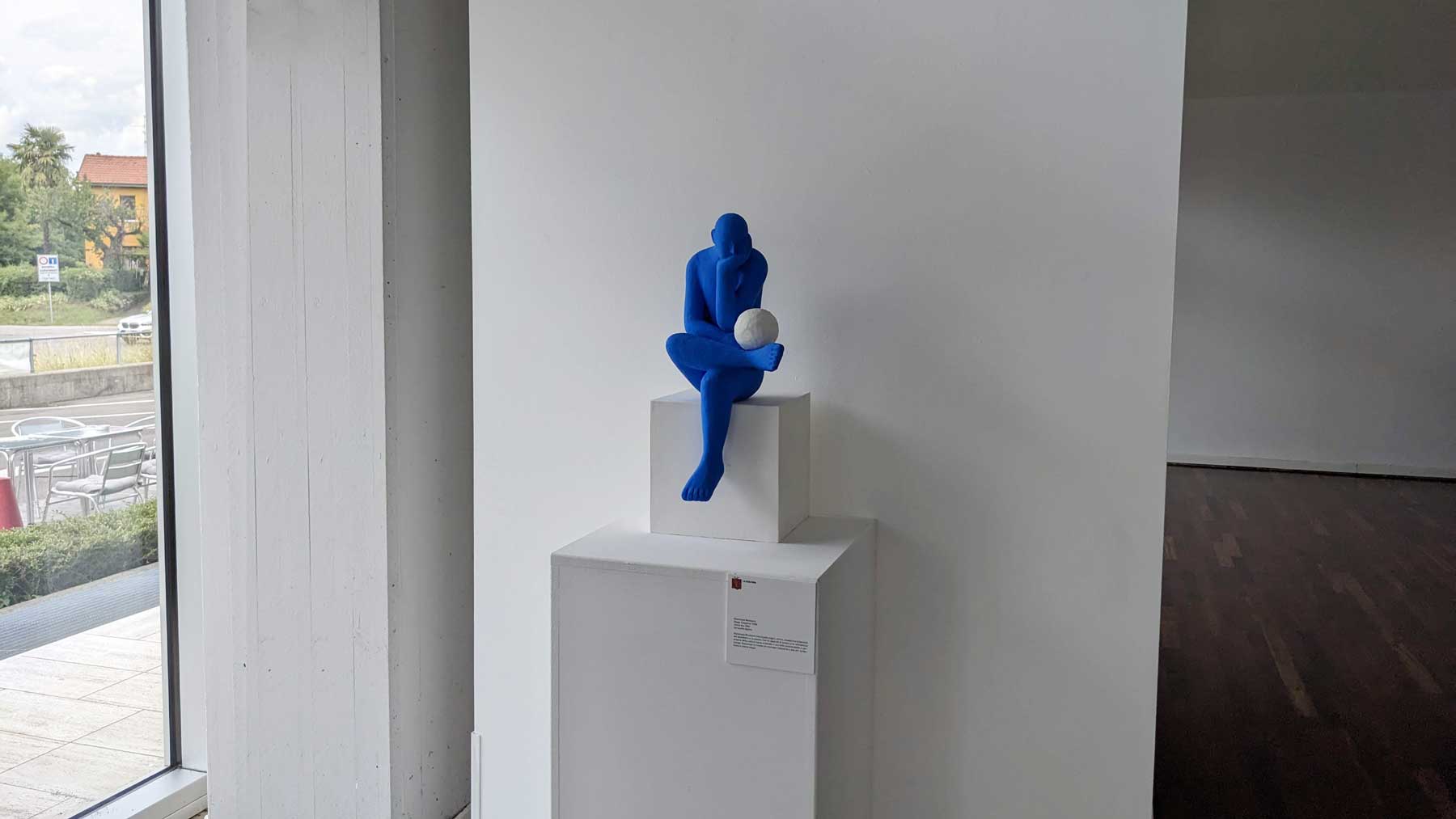 A blue seated human figure looking sideways at a white sphere, Kazumasa Mizokami's 2001 work as it is displayed in this joint exhibition.