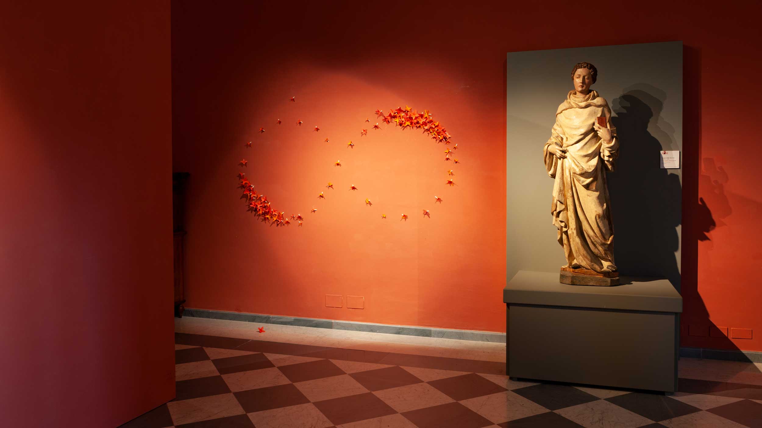 the panorama of the exhibition 2023 Diocesan Museum of Massa, installation work done by kazumasa mizokami title The infinite 2019 250x100x3,5cm plastimol on the red palete movement of the red flowers shape of the infinite. (next to it there is a sculpture with San Leonardo by Jacopo della Quercia, 1420)