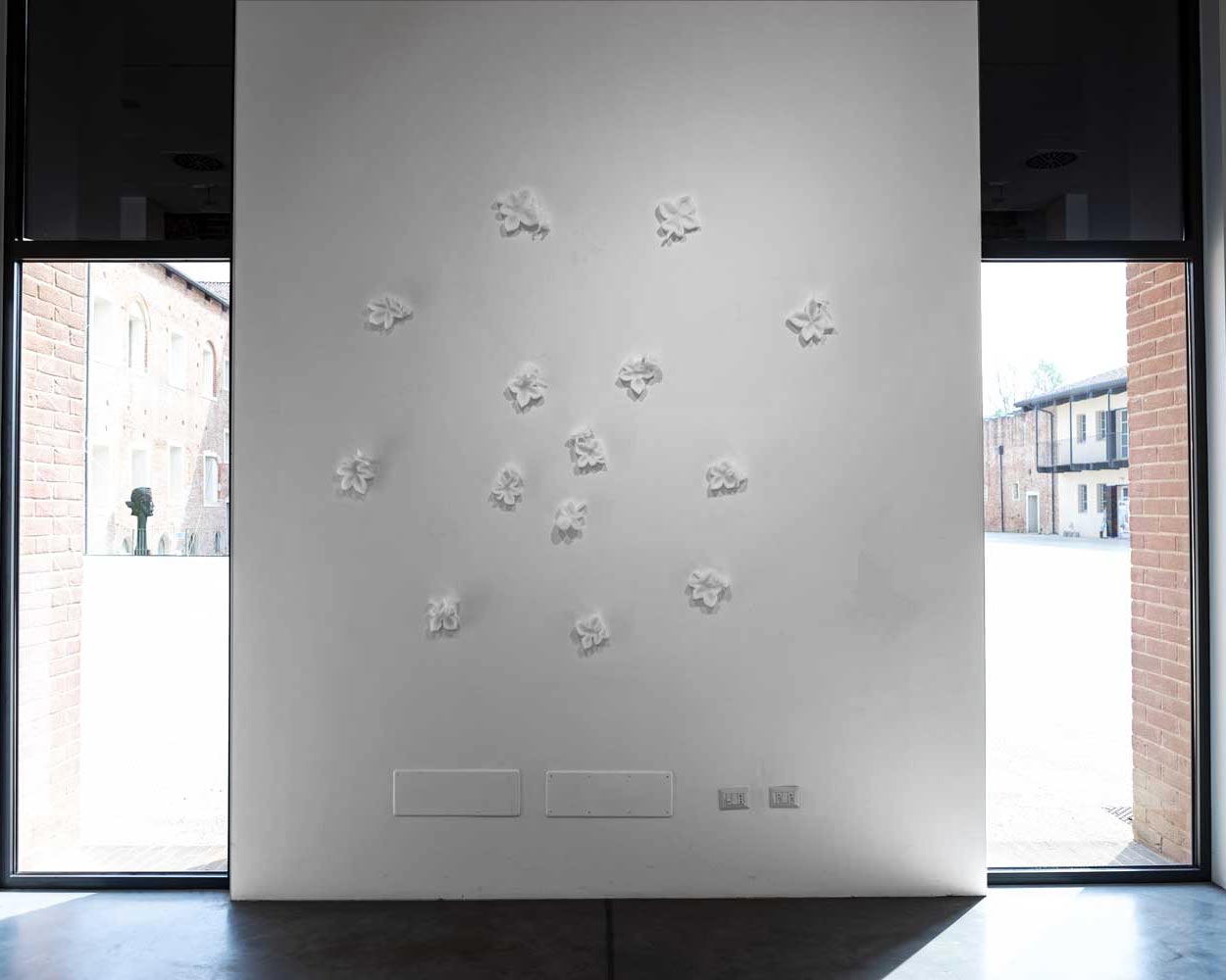 the work "The 12 Living" (2026) by Kazumasa Mizokami is a mural intervention depicting 14 leaves and 12 insects carved in polyurethane foam. These compositions, arranged as a penta circle movement, stand out on a white wall, creating a delicate and subtle contrast. The only distinction between the wall and the works is given by the shadows, which give an ethereal and delicate atmosphere to the whole.