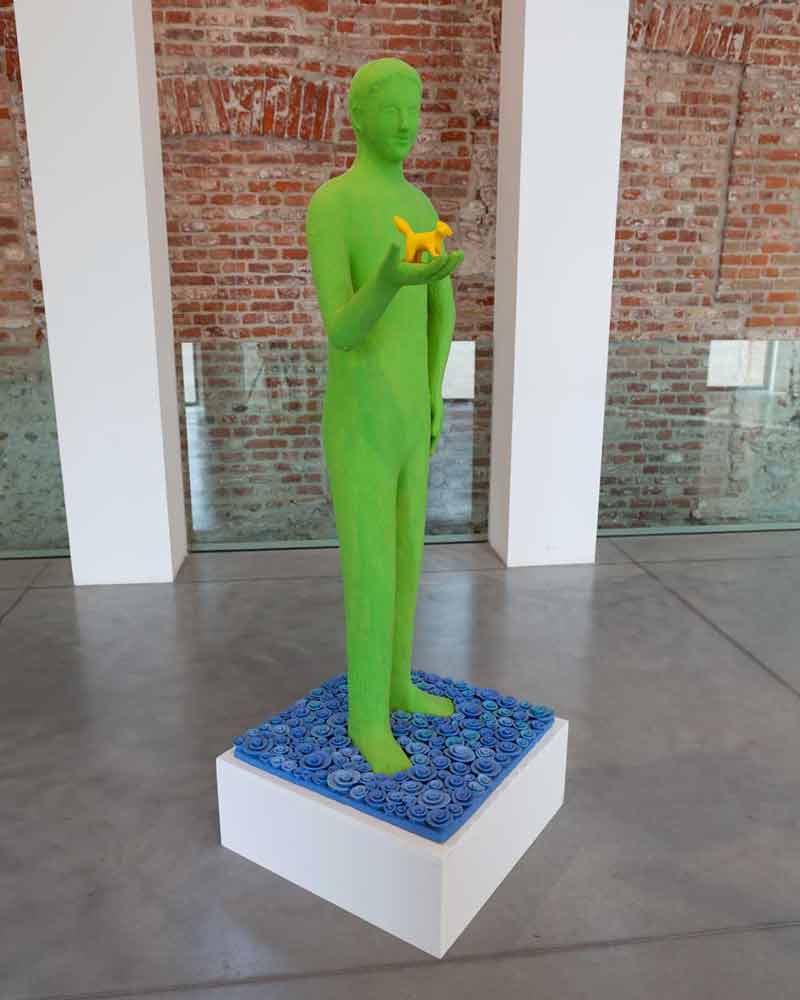 the work "Metamorphosis" (2016) by Kazumasa Mizokami captures attention. This light green statue (162x62x60cm) stands out for its small blue shapes, similar to water waves, applied to the base. On the hand of the statue, a cute little orange animal observes the outside world. The work is made of painted terracotta.