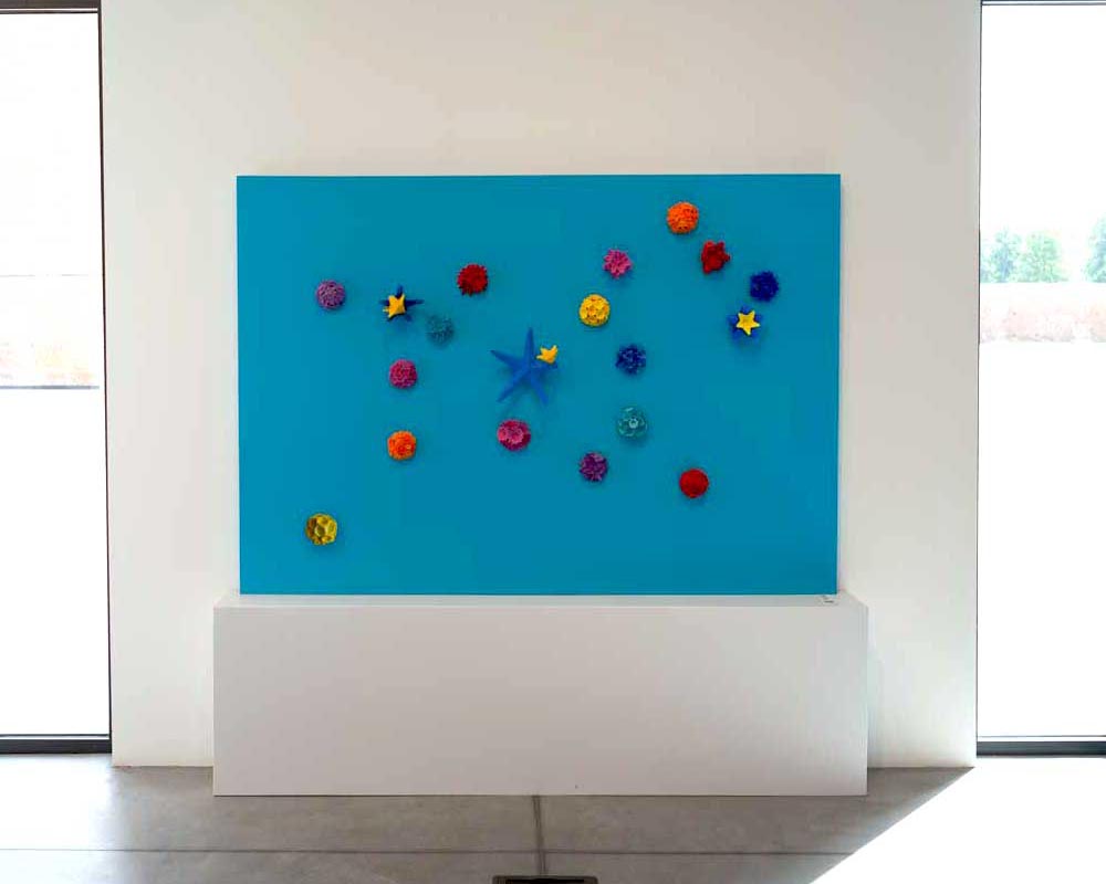 the work "Constellation of lions" by Kazumasa Mizokami is an expressive composition of 19 small colored terracotta objects arranged to form the figure of a lion on a sky blue base of 200x160 cm.